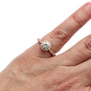 以下が含まれることがあります： ローズゴールドの婚約指輪。中央にラウンドブリリアントカットのダイヤモンドがセットされ、その周りを小さなダイヤモンドが取り囲むハローセッティングが施されています。バンドには、小さなダイヤモンドが敷き詰められた繊細なパヴェセッティングが施されています。