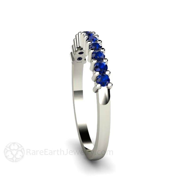 May include: A delicate silver band with a row of blue sapphires set in a curved design. The band is a wedding band or anniversary band.