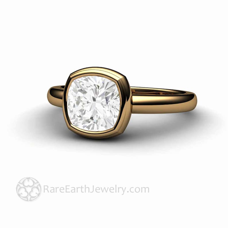 May include: A gold ring featuring a cushion-cut diamond set in a bezel setting. The ring has a thick band and a polished finish.