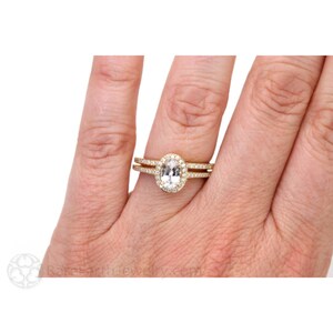 May include: A gold ring set with an oval-shaped gemstone surrounded by a halo of smaller gemstones. The ring is worn on a person's finger.
