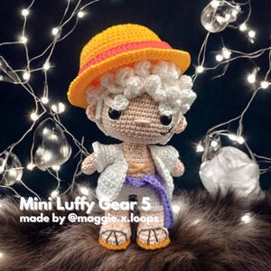 Puede incluir: Una figura de juguete de crochet de Luffy del anime One Piece, con su característico sombrero de paja y una camisa blanca. La figura está en forma de Gear 5, con cabello blanco y un cinturón morado. El juguete está sentado sobre una superficie peluda marrón. "Mini Luffy Gear 5 made by @maggie.x.loops" está escrito debajo de la figura.
