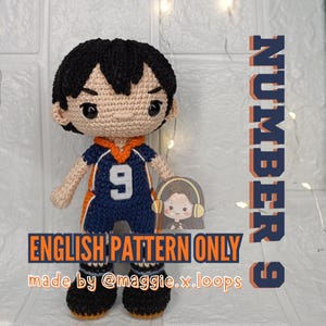 May include: A crocheted doll with black hair and black eyes, wearing a dark blue and orange jersey with the number 9 on the front. The text reads NUMBER 9, ENGLISH PATTERN ONLY, and made by @maggie.x.loops, featuring a small graphic of a person.
