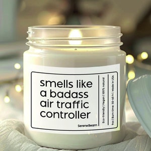May include: A white candle in a glass jar with a black label that reads "smells like a badass air traffic controller". The candle is lit and the label also includes the brand name "Serene Beam" and information about the candle being eco-friendly, vegan, and natural.