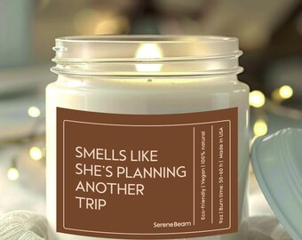May include: A clear glass jar candle with a brown label that reads "Smells like she's planning another trip". The label also includes the text "SereneBeam", "Eco-friendly | Vegan | 100% natural", "9oz | Burn time: 50-60 h | Made in USA".