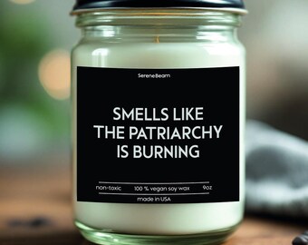 May include: A glass candle jar with a black label that reads "Smells Like The Patriarchy Is Burning". The candle is white and the label also includes the text "non-toxic", "100% vegan soy wax", "made in USA", and "9oz".