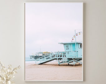 Peut inclure: Une photographie encadrée d'une cabine de sauveteur bleu clair sur une plage de sable avec une jetée en arrière-plan. Le ciel est nuageux et l'eau est bleu clair.
