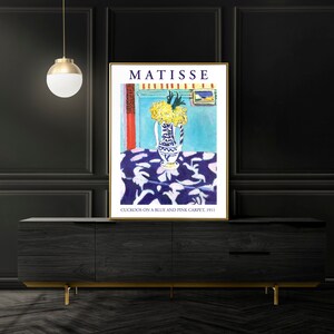 May include: A poster of a painting by Henri Matisse titled "Cuckoos on a Blue and Pink Carpet, 1911". The painting depicts a vase of yellow flowers on a blue and pink patterned tablecloth.