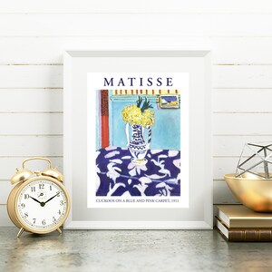 May include: A framed print of a painting by Henri Matisse titled "Cuckoos on a Blue and Pink Carpet, 1911". The painting depicts a vase of yellow flowers on a table with a blue and pink patterned tablecloth.
