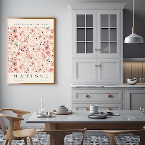 May include: A framed poster with a pink and green floral pattern and the text "PAPIERS DECOUPES MATISSE EDITIONS PARIS FRANCE 1950". The poster is hanging on a white wall in a kitchen with white cabinets and a wooden table.