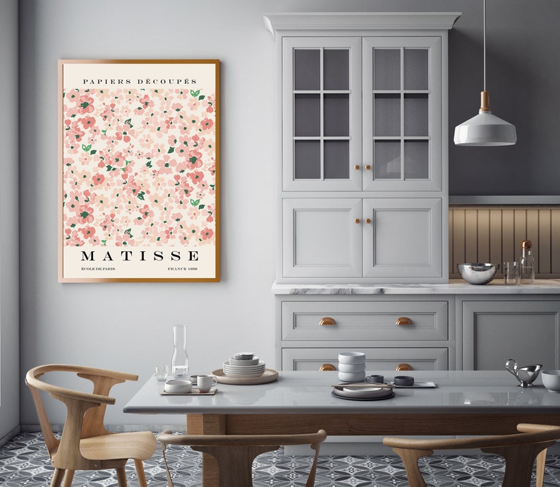 May include: A framed poster with a pink and green floral pattern and the text "PAPIERS DECOUPES MATISSE EDITIONS PARIS FRANCE 1950". The poster is hanging on a white wall in a kitchen with white cabinets and a wooden table.