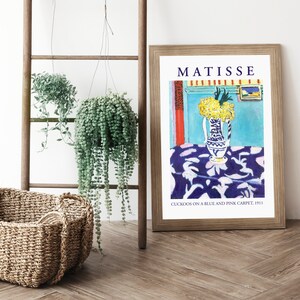 May include: A framed print of a painting by Henri Matisse titled "Cuckoos on a Blue and Pink Carpet, 1911". The painting depicts a vase of yellow flowers on a blue tablecloth with a white and pink pattern.