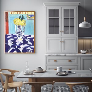 May include: A painting of a still life featuring a blue and white vase filled with yellow flowers on a blue and white patterned tablecloth. The painting is framed in a brown frame and is hanging on a light blue wall.
