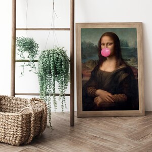 May include: A framed print of the Mona Lisa painting with a pink bubble gum stuck to her mouth. The print is hanging on a white wall next to a wooden ladder with two hanging plants.