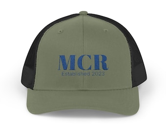 May include: Olive green trucker hat with a black mesh back. The front of the hat features the embroidered text "MCR Established 2023" in blue.