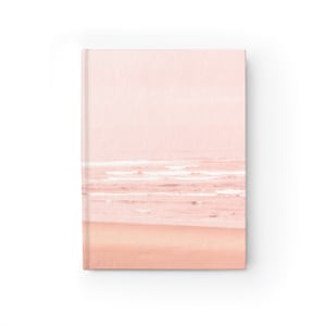 May include: A pink and orange notebook with a beach scene on the cover. The scene features a sandy beach with waves crashing on the shore. The sky is a soft pink color.