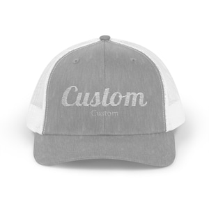 May include: A gray trucker hat with a white mesh back. The word "Custom" is embroidered in a cursive font on the front of the hat. The hat has a curved brim and a button on top.