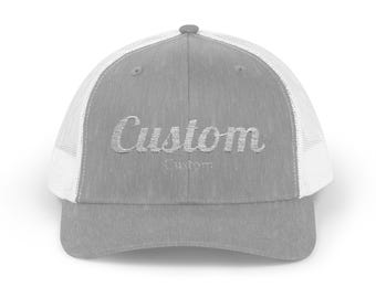 May include: A gray trucker hat with a white mesh back. The word "Custom" is embroidered in a cursive font on the front of the hat. The hat has a curved brim and a button on top.