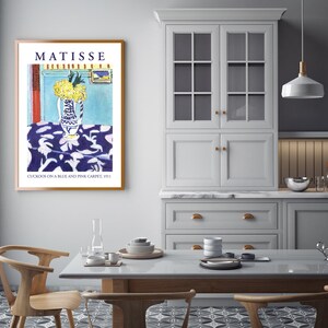 May include: A framed print of a painting by Henri Matisse titled "Cuckoos on a Blue and Pink Carpet, 1911". The painting depicts a vase of yellow flowers on a blue and pink patterned tablecloth.