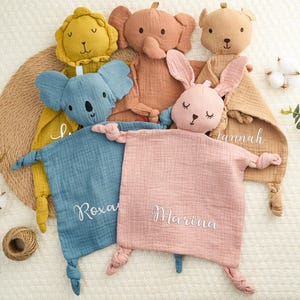 Könnte beinhalten: Vier Plüschtiere mit personalisierten Decken. Die Spielzeuge sind ein Löwe, ein Elefant, ein Koala und ein Hase. Die Decken sind blau, rosa und braun mit den Namen Roxana, Marina und Jannah bestickt.