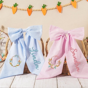 Könnte beinhalten: Zwei Haar-Schleifen zum Thema Ostern, eine hellblaue und eine rosa, mit personalisierten Namen auf den Bändern. Jede Schleife hat einen dekorativen Kranz und befindet sich vor einem festlichen Osterhintergrund mit Karottendekorationen und einem Hasen.