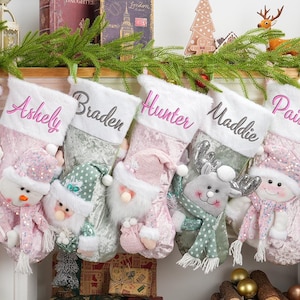 Puede incluir: Cinco calcetines navideños personalizados con nombres bordados en varios colores. Cada calcetín presenta un personaje festivo diferente, incluyendo muñecos de nieve, Papá Noel y un reno, con una textura suave y afelpada. Los calcetines se exhiben en una repisa.