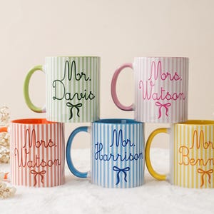 Könnte beinhalten: Fünf Keramiktassen mit vertikalen Streifen in Grün, Pink, Orange, Blau und Gelb. Jede Tasse hat einen farblich passenden Henkel und einen personalisierten Namen in Schreibschrift, darunter Mr. Davis, Mrs. Watson, Mr. Harrison und Mrs. Bennett.