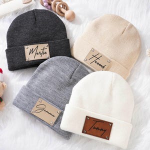 Könnte beinhalten: Vier gestrickte Beanies in verschiedenen Farben mit personalisierten Leder-Patches. Die Beanies sind grau, schwarz, weiß und beige. Die Patches tragen die Namen Martin, Hannah, Gemma und Tommy.