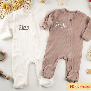 Könnte beinhalten: Zwei Baby-Fußschlafanzüge, einer cremefarben und einer mauvefarben, mit den Namen "Eliza" und "Jude" auf der Brust gestickt. Die Schlafanzüge haben eine gerippte Textur und einen durchgehenden Reißverschluss. Das Bild enthält Babyspielzeug und den Text "FREE Personalization".