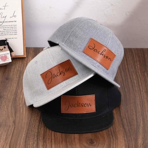 Può includere: Tre cappellini da baseball grigi, grigio chiaro e neri. Ogni cappellino ha una toppa in pelle marrone con il nome "Jackson" in corsivo. I cappellini sono impilati su una superficie di legno.