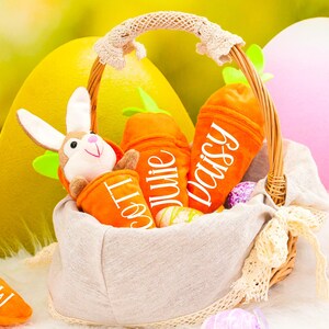 Könnte beinhalten: Ein Weidenkorb gefüllt mit Plüsch-Karottenspielzeug, jedes mit einem bestickten Namen: "Lola", "Julie" und "Daisy". Ein Plüsch-Kaninchen befindet sich ebenfalls im Korb. Der Korb ist von Ostereiern umgeben.