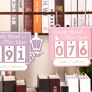 Könnte beinhalten: Zwei hölzerne Bücherzähler, einer in Lavendel und einer in Rosa, jeweils mit einem Laternen-Design. Der lavendelfarbene Zähler zeigt "291" und der rosa Zähler "076". Beide zeigen "Gelesene Bücher dieses Jahr" und "Jodie's Library".