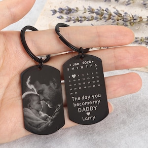 Könnte beinhalten: Zwei schwarze Schlüsselanhänger im Dog-Tag-Design. Einer zeigt ein Graustufenbild von Vater und Baby. Der andere zeigt einen Kalender für Januar 2026 und den Text "The day you become my DADDY" mit einem Namen.