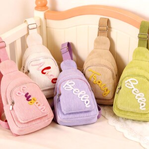 Könnte beinhalten: Fünf Cord-Umhängetaschen in Pastellfarben: Rosa, Weiß, Lila, Beige und Hellgrün. Jede Tasche hat eine kleine Vordertasche und einen gestickten Namen: "Sweet", "Mia", "Bella", "Tracy" und "Bella".