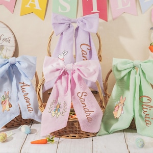 Könnte beinhalten: Drei österliche Schleifendekorationen in Pastellfarben: Blau, Rosa und Grün. Jede Schleife hat einen personalisierten Namen auf dem Band und eine Hasenillustration. Die Schleifen werden in Weidenkörben präsentiert.