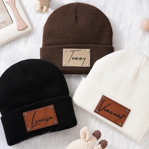 Könnte beinhalten: Drei gestrickte Beanies mit personalisierten Leder-Patches. Die Beanies sind braun, schwarz und weiß. Die Patches haben die Namen "Tommy", "Vincent" und "Louise" aufgedruckt.