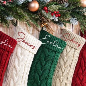 May include: Five cable-knit Christmas stockings in red, cream, green, and beige, each with a name embroidered in a complementary color. Festive greenery and ornaments adorn the top, creating a warm holiday scene.