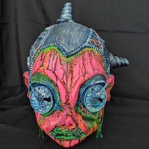 May include: A pink and green face mask with blue and silver details. The mask has a textured surface and appears to be made of a hard material. The mask has a unique and slightly disturbing design.