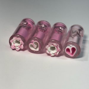 May include: Four clear glass stampers with pink and white floral and heart designs. Each stamper has a cylindrical shape with a pink interior and a decorative end featuring either a paper flower or a heart shape.