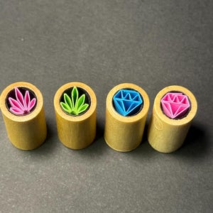 May include: Four wooden cylindrical stamps with colorful designs. The designs include a pink flower, a green leaf, a blue diamond, and a pink diamond. The stamps are arranged in a row against a dark background.