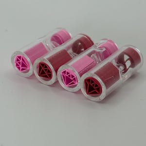 Puede incluir: Cuatro tubos transparentes de brillo labial, con tonos rosas y rojos. Cada tubo presenta un diseño en forma de diamante rosa en la base. Los colores del brillo labial varían de rosa claro a rojo intenso.