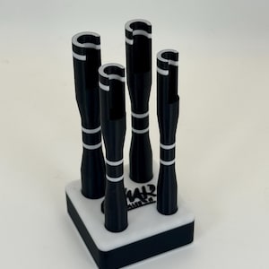 May include: A black and white pen holder with five pens. The pens are black with white stripes and a white top. The holder is a white square with a black border. The text "WAR" is visible on the holder.