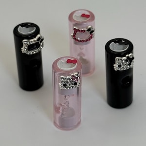 May include: Four lipstick tubes, two black and two pink, with decorative Hello Kitty embellishments. The black tubes have silver rhinestone accents, while the pink tubes feature pink rhinestone accents. The lipstick tubes are cylindrical and stand upright.