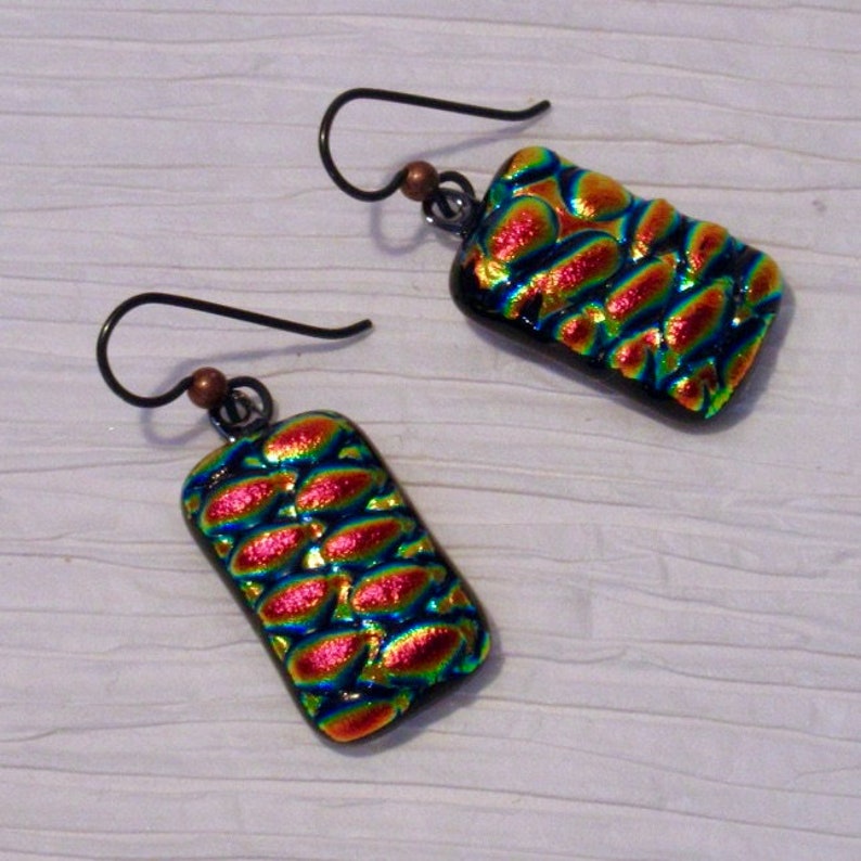 May include: A pair of iridescent glass earrings with a textured pattern. The earrings are rectangular in shape and have a black background with red, green, and blue iridescent colors. The earrings are attached to silver hooks.