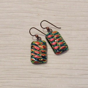 May include: A pair of iridescent earrings with a copper wire and a rectangular glass pendant. The glass pendant has a textured pattern of overlapping circles in shades of green, blue, and pink.