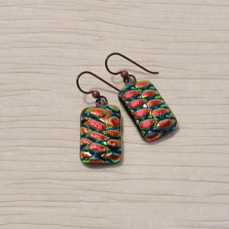 May include: A pair of iridescent earrings with a copper wire and a rectangular glass pendant. The glass pendant has a textured pattern of overlapping circles in shades of green, blue, and pink.