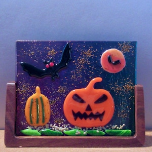 May include: A glass art piece depicting a Halloween scene with a black bat flying over a purple and gold background with a pumpkin and a smaller pumpkin. The pumpkin has a scary face and the background has a glittery texture.