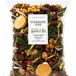 May include: A clear plastic bag filled with Evermere Pine handcrafted potpourri. The contents include brown pinecones, green cedar sprigs, round wooden slices, dried berries, and small tan spheres. A white label features the text Evermere Pine.