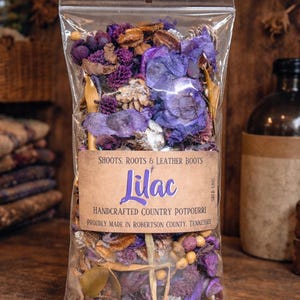 May include: A clear plastic bag filled with dried lilac potpourri. The bag features a brown label with the word "Lilac" and the text "Handcrafted Country Potpourri". The potpourri includes purple, brown, and beige dried flowers and botanicals.