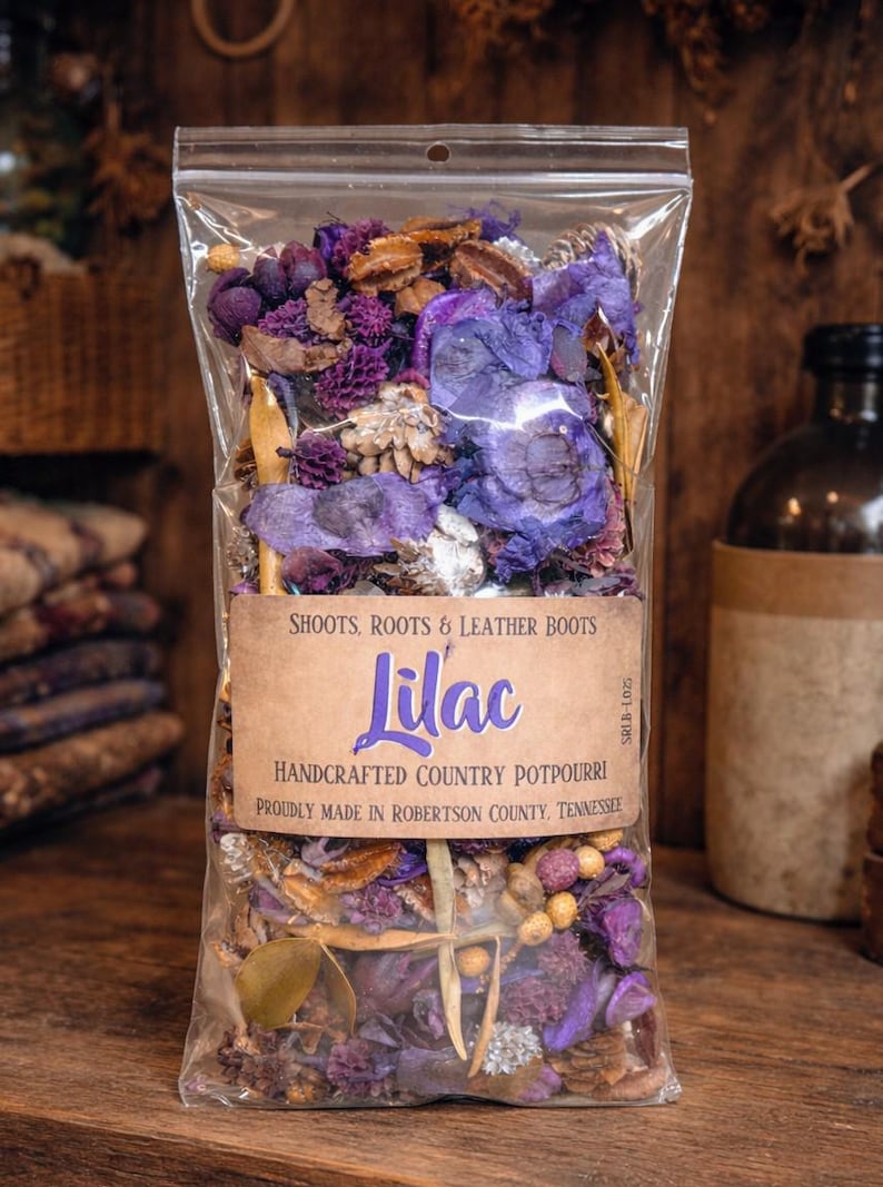 May include: A clear plastic bag filled with dried lilac potpourri. The bag features a brown label with the word "Lilac" and the text "Handcrafted Country Potpourri". The potpourri includes purple, brown, and beige dried flowers and botanicals.