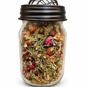 May include: A clear glass jar filled with dried herbs, flowers, and berries. The jar has a black metal lid and a decorative wire mesh top. The contents include green, brown, and pink elements, suggesting a potpourri or herbal blend.